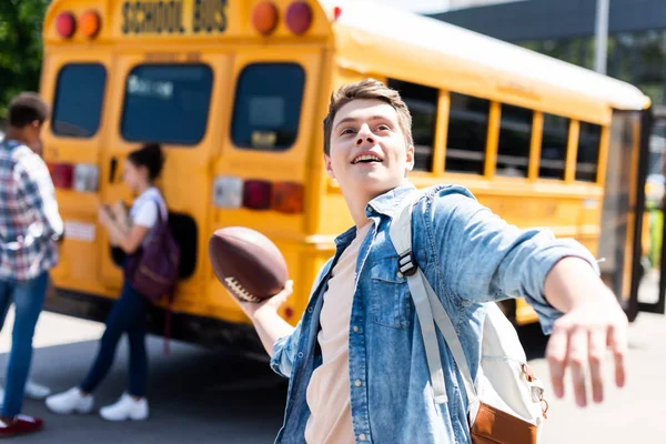 Smiling teen schoolboy throwing american football ball in front of school bus — Stock Photo