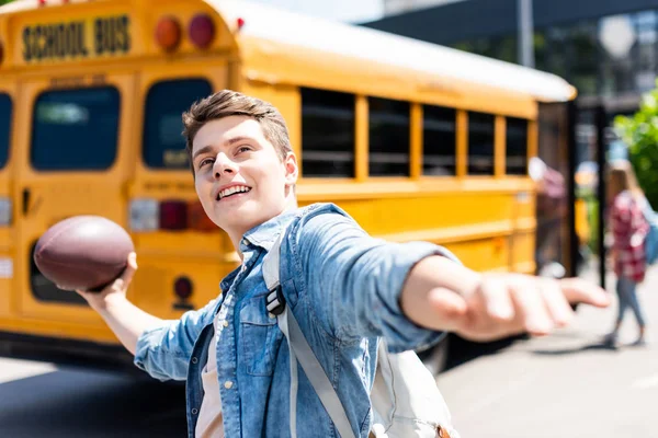 Feliz adolescente escolar lanzando americano pelota de fútbol en frente de autobús escolar - foto de stock