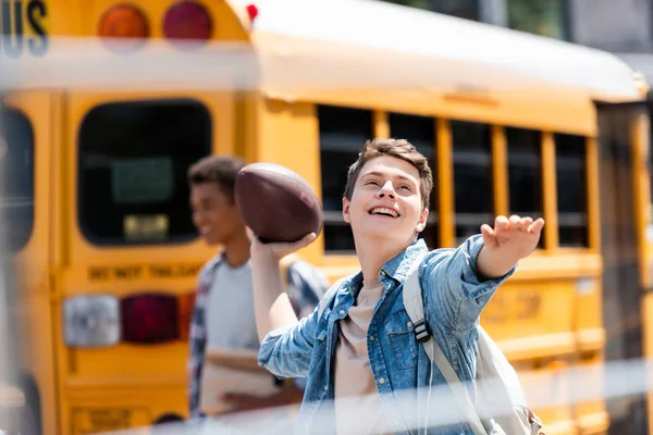 Feliz adolescente escolar lanzando americano pelota de fútbol en frente de la escuela autobús con borrosa compañero de clase caminando sobre fondo - foto de stock