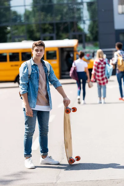 Happy teen schoolboy with skateboard standing in front of school bus and group of classmates walking blurred on background — Stock Photo