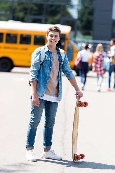 Happy teen schoolboy with skateboard standing in front of school bus — Stock Photo