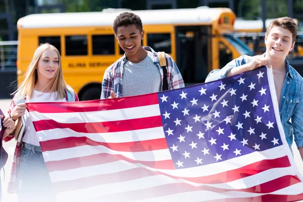 Groupe d'adolescents souriants tenant le drapeau des Etats-Unis devant le bus scolaire — Photo de stock