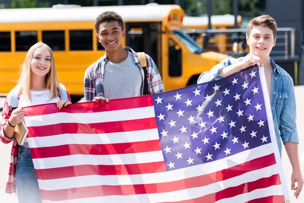 Grupo de estudiantes adolescentes felices sosteniendo bandera de EE.UU. frente al autobús escolar - foto de stock