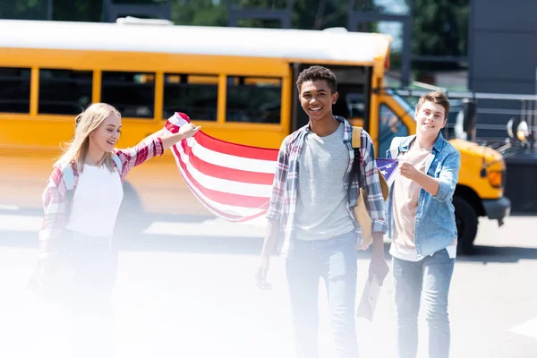 Grupo de estudiantes adolescentes felices con bandera de EE.UU. frente al autobús escolar - foto de stock