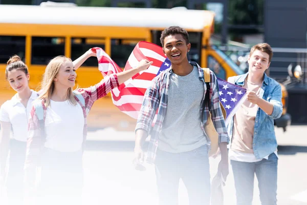 Grupo de eruditos adolescentes multiétnicos americanos caminando con bandera de EE.UU. frente al autobús escolar - foto de stock