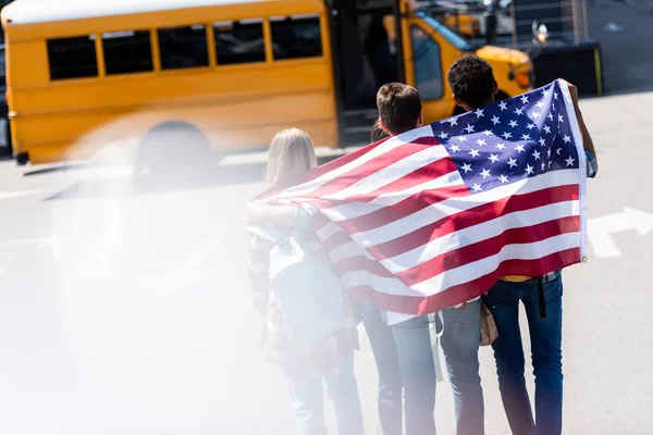 Rear view of group of american teen scholars with usa flag in front of school bus — Stock Photo