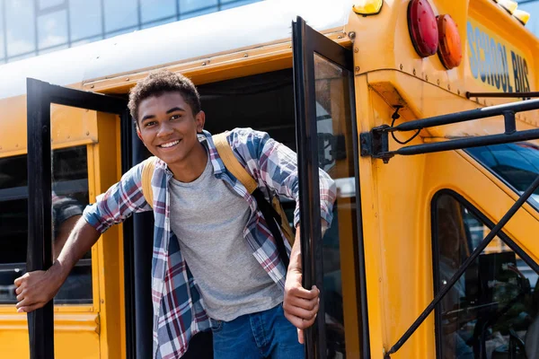 Adolescente feliz afroamericano colegial de pie en la puerta del autobús escolar y mirando a la cámara - foto de stock
