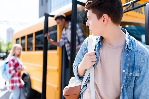 Teen schoolboy walking in front of school bus and turning back at classmates — Stock Photo