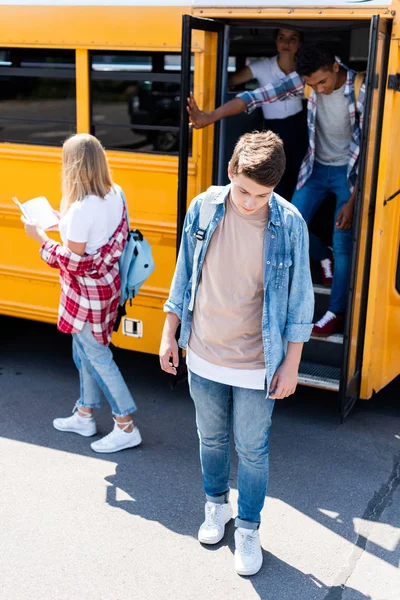 Sad teenager schoolboy standing in front of school bus with classmates — Stock Photo