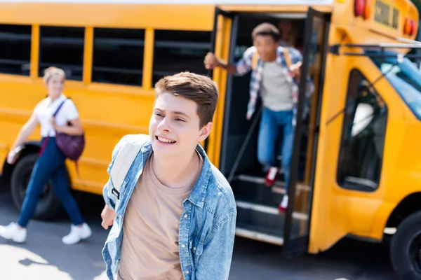 Feliz adolescente escolar corriendo fuera de la escuela autobús con borrosa compañeros de clase en el fondo - foto de stock