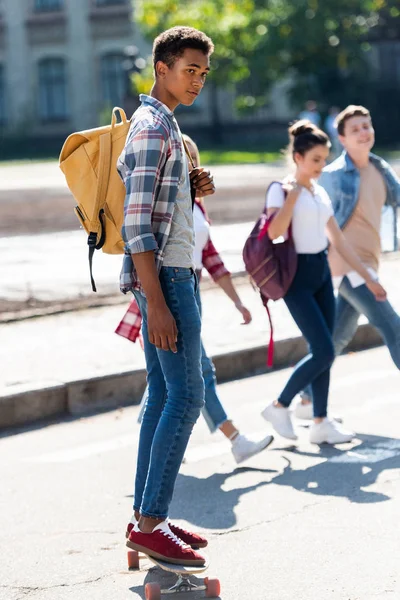Joven africano americano colegial montar monopatín con borrosa compañeros de clase caminando sobre fondo - foto de stock