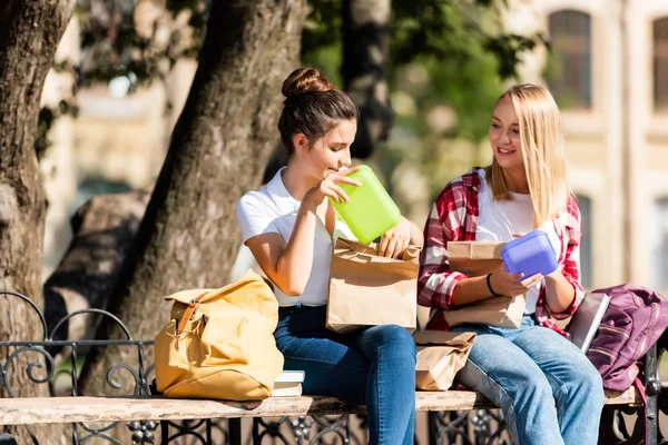 Happy teen schoolgirls sitting on bench with lunch boxes — Stock Photo