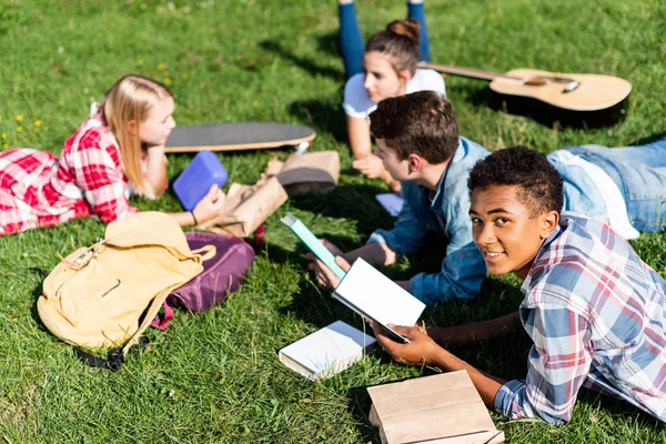 Groupe de chercheurs adolescents multiethniques couchés sur l'herbe et étudier ensemble — Photo de stock