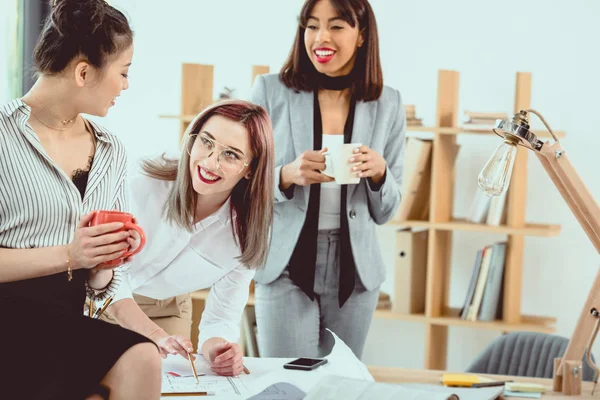 Multiethnic group of young businesswomen working with blueprint and drinking coffee in office — Stock Photo