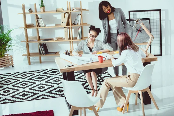Lächelnde junge Geschäftsfrauen, die mit Entwürfen in modernen Büros arbeiten — Stockfoto