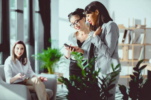 Smiling young businesswomen standing together and using smartphone while colleague sitting behind — Stock Photo