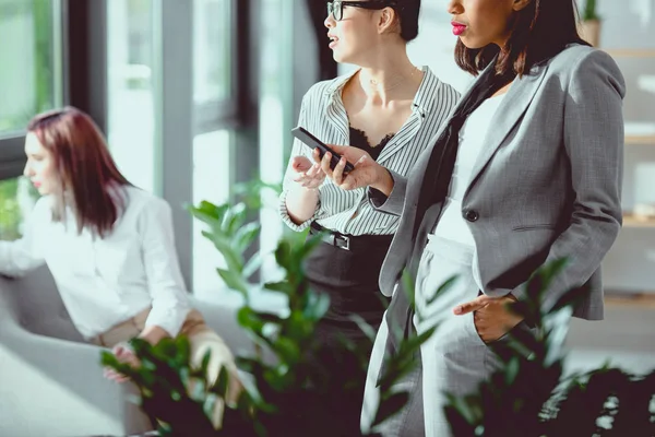 Jeunes femmes d'affaires debout et utilisant un smartphone avec un collègue assis derrière — Photo de stock