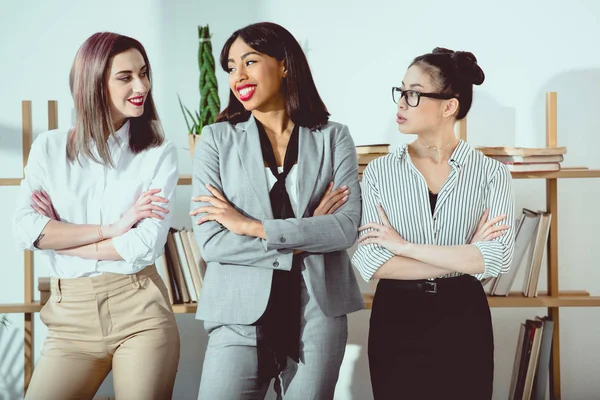 Mujeres de negocios multiétnicas sonrientes en ropa formal de pie con los brazos cruzados - foto de stock