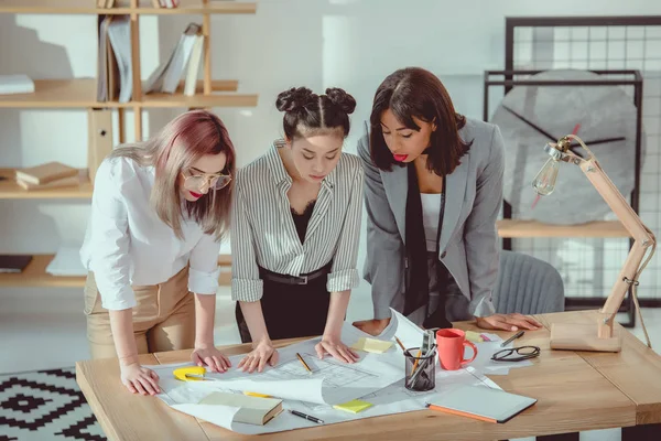 Jóvenes diseñadores multiétnicos mirando los planos mientras están de pie en la oficina - foto de stock