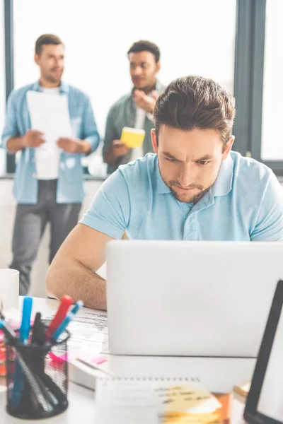 Концентрированный молодой бизнесмен с помощью ноутбука в то время как коллеги стоят позади в офисе — стоковое фото