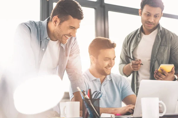 Jóvenes empresarios multiétnicos sonrientes usando laptop y tomando notas en la oficina - foto de stock