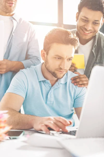 Ориентированный молодой бизнесмен с помощью ноутбука и коллег, стоящих позади в офисе — стоковое фото