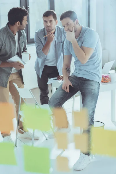 Jeunes hommes d'affaires multiethniques concentrés regardant des notes collantes tout en travaillant à un nouveau projet ensemble — Photo de stock
