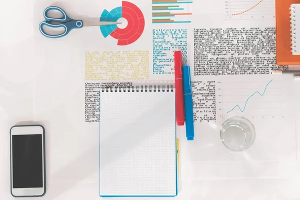 Vista superior del smartphone con pantalla en blanco, cuaderno vacío con marcadores, tijeras y gráficos de negocios en el lugar de trabajo - foto de stock