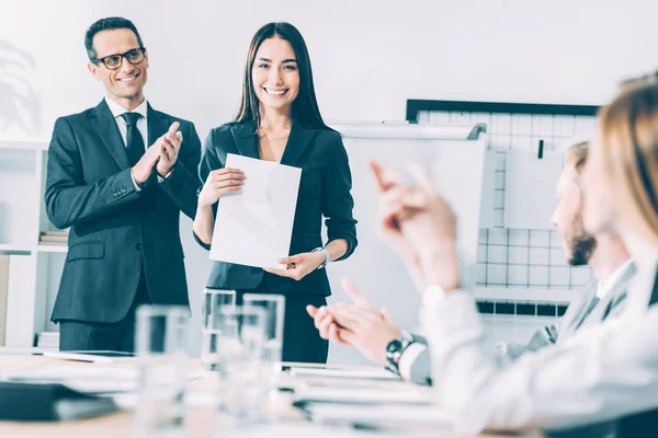 Rewarded happy asian manageress holding blank paper while colleagues clapping to her — Stock Photo