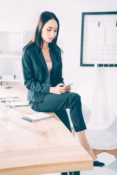 Asiático mujer de negocios usando smartphone mientras sentado en mesa en oficina — Stock Photo