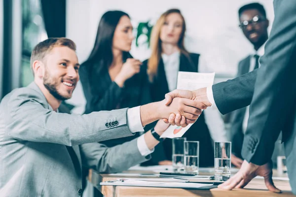 Socios comerciales estrechando la mano con colegas difusos multiculturales en el fondo - foto de stock