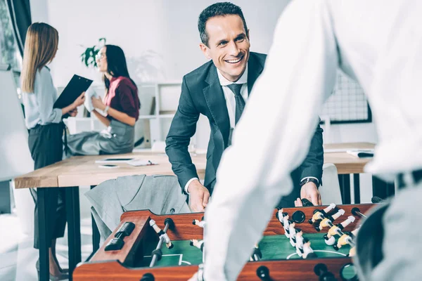 Homens de negócios jogando futebol de mesa no escritório moderno com mulheres de negócios falando borrado no fundo — Fotografia de Stock