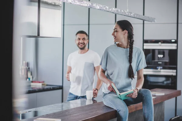 Novia sonriente sentada en el mostrador de la cocina con libro abierto, novio sosteniendo tazas de café - foto de stock