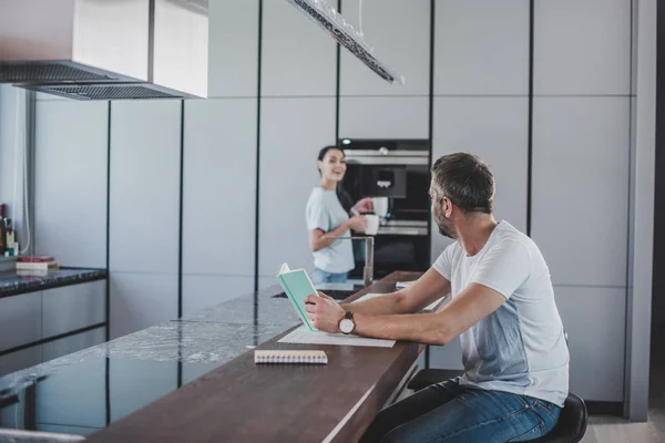 Vista lateral do namorado segurando livro e namorada de pé com xícaras de café na cozinha — Fotografia de Stock