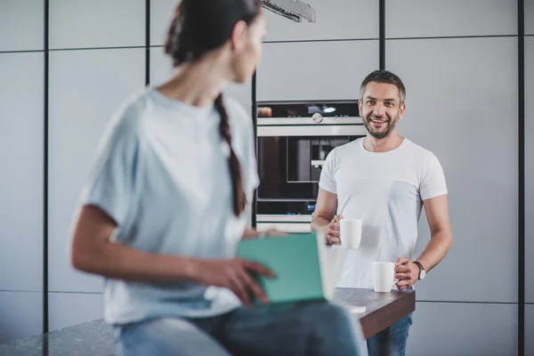 Girlfriend sitting on kitchen counter with open book and looking at boyfriend with coffee cups — Stock Photo