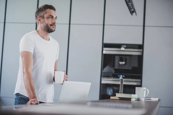 Hombre guapo sosteniendo la taza de café y mirando hacia otro lado en la cocina - foto de stock