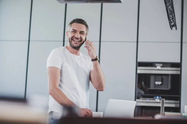 Smiling handsome man talking by smartphone in kitchen — Stock Photo