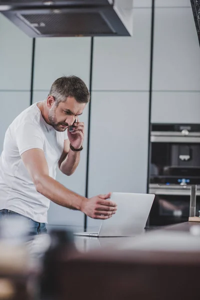 Handsome freelancer talking by smartphone and looking at laptop in kitchen — Stock Photo