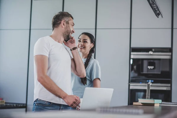 Smiling boyfriend talking by smartphone and looking at girlfriend in kitchen — Stock Photo