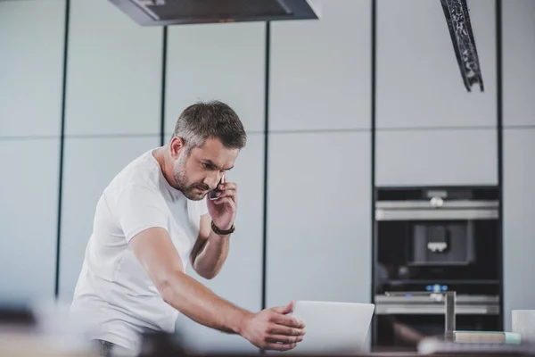 Vista lateral del hombre guapo hablando por teléfono inteligente y mirando el ordenador portátil en la cocina - foto de stock