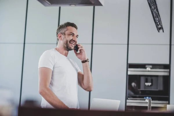 Улыбающийся красивый мужчина разговаривает со смартфоном на кухне и смотрит в сторону — стоковое фото