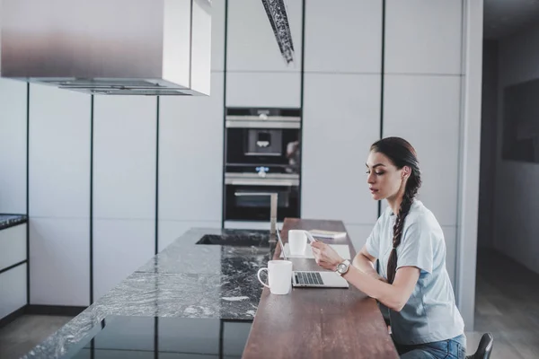Seitenansicht der schönen Freiberuflerin, die am Laptop arbeitet und das Smartphone in der Küche hält — Stockfoto