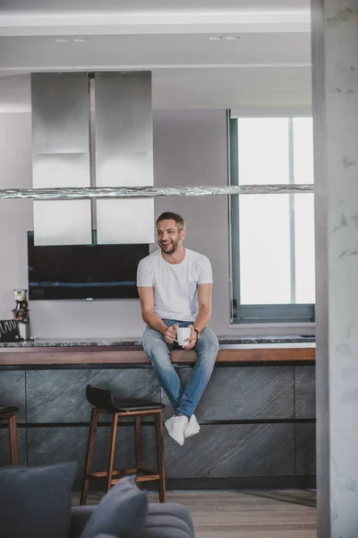 Hombre guapo sonriente sentado en el mostrador de la cocina con taza de café en la mañana - foto de stock