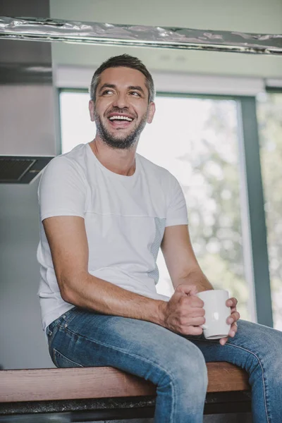 Smiling handsome man sitting on kitchen counter with cup of tea in morning and looking away — Stock Photo