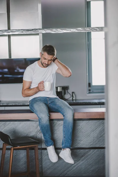 Handsome man sitting on kitchen counter and looking in cup in morning — Stock Photo