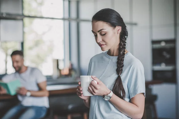 Selective focus of attractive woman standing with coffee cup while her boyfriend reading book behind in kitchen at home — Stock Photo