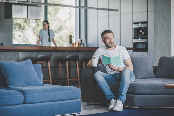 Erwachsener Mann mit Kaffeetasse liest Buch auf Sofa, während seine Freundin zu Hause in der Küche steht — Stockfoto