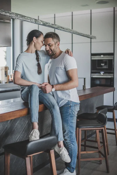 Mujer sonriente sentada en el mostrador de la cocina y abrazando novio en casa - foto de stock