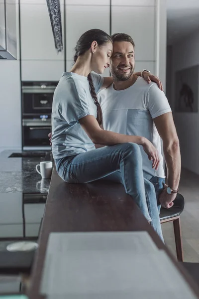 Vista lateral de la atractiva mujer sentada en el mostrador de la cocina y abrazando novio en casa - foto de stock
