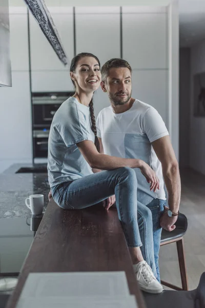Щаслива молода жінка сидить на кухонній стійці, а її хлопець стоїть поруч вдома — стокове фото